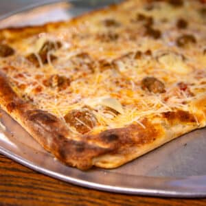 Close-up of a rectangular deep-dish pizza slice topped with melted mozzarella cheese, meatballs, and Parmesan on a golden, crispy crust served on a silver tray.