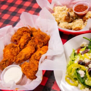 Basket of crispy buffalo chicken wings served with ranch dressing, alongside a plate of fresh Greek salad and a basket of garlic knots with marinara sauce on a red-and-black checkered tablecloth.
