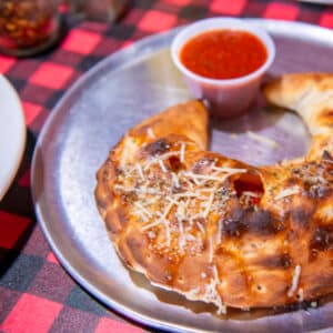 Freshly baked calzone topped with melted cheese and herbs, served on a metal plate with a side cup of marinara dipping sauce on a red-and-black checkered tablecloth.