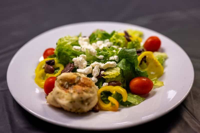 A Greek salad with feta, cherry tomatoes, olives and banana peppers beside a baked knot on a white plate